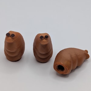 May include: Three brown, worm-like figurines with dark eyes. Two stand upright, while the third lies on its side, revealing a hollow opening. The figurines have a smooth, rounded appearance.