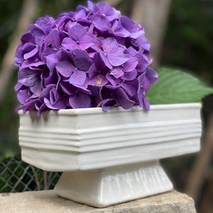 Puede incluir: Un jarrón rectangular de cerámica blanca con base de pedestal contiene un gran racimo de vibrantes flores de hortensia moradas. Una hoja verde asoma por detrás de las flores. El jarrón está sobre una superficie de piedra.
