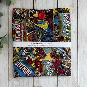 May include: A padded fabric book sleeve with a comic book print featuring characters such as Spider-Man, Iron Man, and Wolverine. The sleeve has a red button closure and a white label that reads "Padded Fabric Book Sleeve Handmade by Claire Michele Designs".