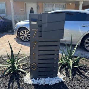 May include: A dark gray vertical mailbox featuring horizontal slats and the house number 7710 displayed in gold-colored numerals. The mailbox includes a rectangular top compartment and a small solar light fixture positioned above the house numbers.