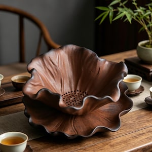 Puede incluir: Un colador de té de cerámica marrón con forma de flor de loto, con una base a juego. El colador tiene una serie de pequeños agujeros en el centro. El juego de té está sobre una mesa de madera.