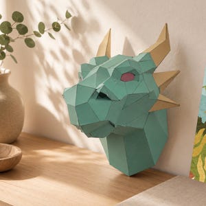 Peut inclure: Une tête de dragon en papier turquoise avec des cornes dorées et des yeux roses est fixée au mur. La sculpture géométrique est exposée sur une surface en bois, à côté d'un vase avec de la verdure et une impression décorative.