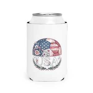 May include: A white can cooler featuring a graphic of a soccer ball decorated with red, white, and blue floral patterns and stars. The text USA is printed in a bold serif font below the ball, framed by a decorative wreath of green leaves.
