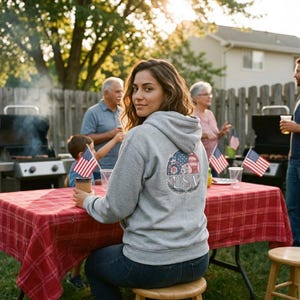 May include: A light gray hooded sweatshirt featuring a circular graphic on the back. The graphic contains a patchwork pattern of stars and stripes in red, white, and blue, with the text USA centered below the pattern, framed by leafy branches.