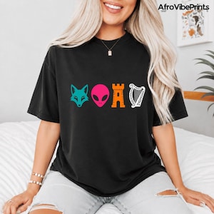 May include: Black t-shirt with a colourful graphic design featuring a teal fox head, a pink alien head, an orange castle, and a white harp. The shirt has a comfortable, oversized fit.