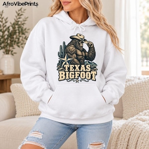 May include: A white hooded sweatshirt featuring a graphic of a brown Bigfoot wearing a cowboy hat, standing next to a cactus and a star. The text "TEXAS BIGFOOT" is printed in large, bold, tan-colored letters below the illustration.