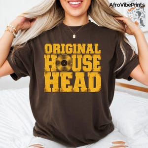 May include: A dark brown short-sleeve t-shirt featuring the yellow text ORIGINAL HOUSE HEAD printed on the front. The letter O in the word HOUSE is replaced by a graphic illustration of a black vinyl record with a gold center label.