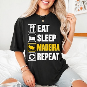 Puede incluir: Una camiseta negra de manga corta con un estampado gráfico en blanco y amarillo. El diseño incluye cuatro filas apiladas con iconos y texto: un tenedor y una cuchara con EAT, una cama con SLEEP, un mapa de Madeira con MADEIRA y flechas con REPEAT.
