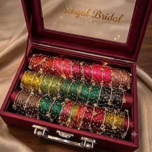 May include: A maroon velvet jewellery box with a clear lid featuring the gold text Sehgal Bridal. Inside, three rows of thin, multicoloured bangles are arranged on velvet cushions. The bangles are metallic, featuring red, green, yellow, and brown hues with small gold-toned bead accents.