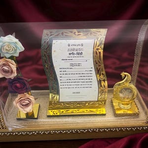 Puede incluir: Una vitrina decorativa con cubierta arqueada transparente, bordes dorados y detalles de espejo. En el interior, una placa central con marco dorado y texto en punjabi está flanqueada por cuatro rosas artificiales y una figura de pavo real.