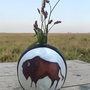 May include: A spherical ceramic vase with a white circular panel featuring a brown painted bison. The vase has a black rim and base, and contains a small bouquet of slender green stems with tiny purple flower buds at the top.