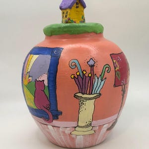 May include: A round, peach-colored ceramic jar with a green lid topped by a small yellow house with a purple roof. The jar features hand-painted illustrations of a pink cat looking out a blue window, a vase of colorful umbrellas, and a red lamp.