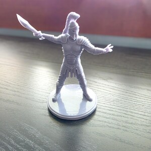 May include: A gray miniature gladiator figurine, posed with a sword held high. The figurine is wearing armor and a helmet, standing on a circular base. The figure is a detailed representation of a warrior, suitable for tabletop gaming or display.