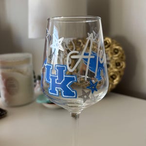 May include: A clear glass wine glass featuring hand-painted designs in blue and white. The glass displays the letters UK in a block font, the name Alex in cursive script, and several star shapes scattered around the exterior surface.