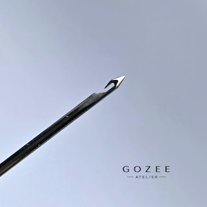 Op de afbeelding: Een zilverkleurige metalen naald met een scherpe punt en een duidelijk open haakmechanisme aan het uiteinde. De naald heeft een glad, reflecterend oppervlak. De tekst GOZEE ATELIER staat in het zwart in de rechterbenedenhoek gedrukt.