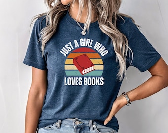 Può includere: Una maglietta blu navy con una grafica in stile vintage che dice "Just a girl who loves books" e presenta un libro rosso su sfondo di tramonto.