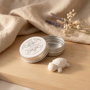 Könnte beinhalten: Eine runde silberne Metalldose mit einem Deckel, der eine Schildkrötengrafik und den Text JULIE LOVES ROMEO SCENTI DUFT-SCHILDKRÖTE LIGHT SCENTS RITUALS zeigt. Neben der offenen Dose liegt ein kleines weißes Objekt in Schildkrötenform.