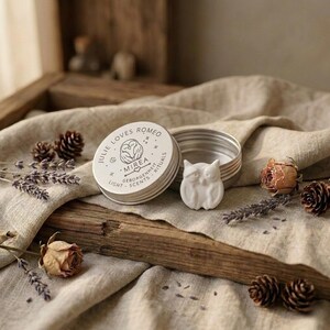 Könnte beinhalten: Eine runde silberne Metalldose mit einem Deckel, der eine Eulenillustration und den Text JULIE LOVES ROMEO MIREA GEBORGENHEIT LIGHT SCENTS RITUALS zeigt. Neben der offenen Dose liegt ein kleines weißes eulenförmiges Objekt, umgeben von getrocknetem Lavendel und Tannenzapfen.