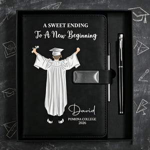 Puede incluir: Un cuaderno negro con un cierre metálico plateado y un bolígrafo negro a juego en una caja de regalo. La portada presenta una ilustración blanca de un graduado con toga y birrete, con el texto A SWEET ENDING To A New Beginning, David, POMONA COLLEGE 2026.