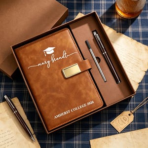 Puede incluir: Un juego de diario y bolígrafos de cuero marrón en una caja de regalo. El diario está grabado con un birrete, el nombre "Mary Brandt" y "AMHERST COLLEGE 2026". El juego incluye dos bolígrafos, uno plateado y otro marrón.