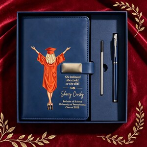 Puede incluir: Un cuaderno azul marino y un bolígrafo en una caja de regalo. La portada muestra a una graduada rubia con toga naranja y brazos levantados, el texto "She believed she could, so she did!", el nombre "Sherry Crosby" y "Bachelor of Science University of Pennsylvania Class of 2025".