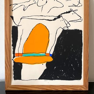 Peut inclure: Une impression encadrée présentant un dessin au trait noir stylisé d'une vache debout sur une grande forme orange avec une bande turquoise et rouge. Le fond est divisé entre crème et noir, avec des croquis d'animaux au-dessus dans un cadre en bois.