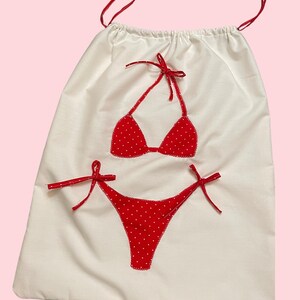 May include: A white drawstring bag with a red polka dot bikini applique. The bikini top and bottom are detailed with red bows. The bag has red drawstring cords.