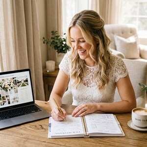May include: A spiral-bound wedding planner book with white pages and black text, open on a wooden table. Next to it sits a laptop displaying a photo collage of wedding flowers, a ceramic mug with text, and a glass vase filled with white flowers.