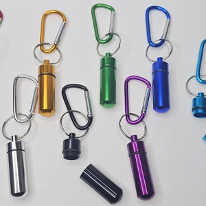 May include: A collection of cylindrical metal pill containers in red, gold, green, blue, silver, black, and purple. Each container features a matching carabiner attached by a split ring. Some containers are shown open, revealing the hollow interior.