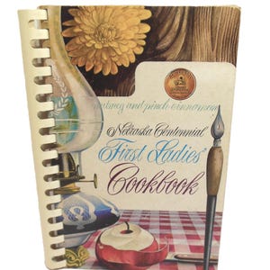 May include: A spiral-bound cookbook titled Nebraska Centennial First Ladies Cookbook with a cover illustration featuring a yellow flower, a glass oil lamp, a red and white checkered tablecloth, a sliced apple, and a fountain pen with a gold seal.