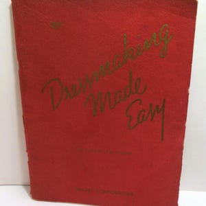 May include: A vintage red book titled "Dressmaking Made Easy" with gold lettering. The book is an American edition by McCall Corporation. The cover has a textured appearance and shows signs of wear.