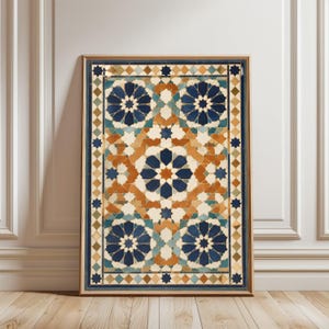 May include: Framed artwork with a geometric pattern in blue, orange, and cream. The design features star and floral motifs, framed in a wooden border. The artwork is displayed against a white wall and wooden floor.