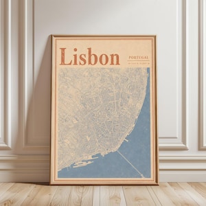May include: Framed vintage-style map print of Lisbon, Portugal. The map features a detailed blue and beige city layout, with the word "Lisbon" in large, rust-colored letters at the top. Coordinates are also included. The print is in a wooden frame.