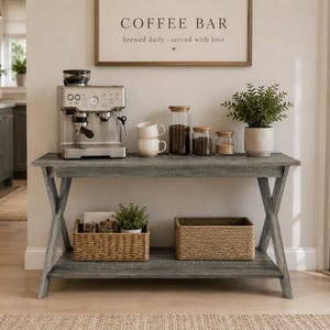 May include: A gray wooden console table with an X-frame design and a lower shelf. The top holds a silver espresso machine, two stacked white mugs, three glass canisters, and a potted plant. The lower shelf contains two woven baskets and a plant.