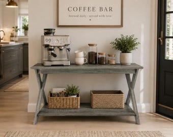 May include: A gray wooden console table with an X-frame design and a lower shelf. The top holds a silver espresso machine, two stacked white mugs, three glass canisters, and a potted plant. The lower shelf contains two woven baskets and a plant.