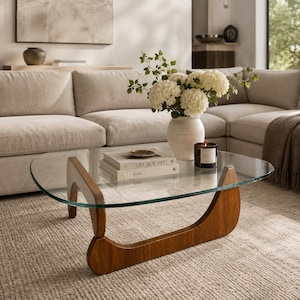 May include: A coffee table with a clear, curved glass top and a solid wood base featuring two arched, interconnected supports. On the glass surface sits a white ceramic vase filled with white hydrangea flowers and a black candle in a glass jar.
