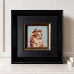 May include: A square framed painting featuring a portrait of a long-haired orange cat against a light blue background. The artwork is encased in a thick black frame with an inner gold-toned decorative border, creating a high-contrast display.