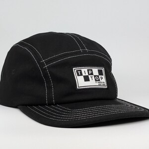 May include: Black five-panel cap with white stitching and a flat peak. The cap has a rectangular patch with the words "TIP TOP" in a checkerboard design. The text "TT HATS CO. EST 1986" is below the logo.