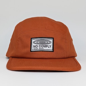 May include: A rust-coloured, five-panel cap with a flat peak. The cap has a rectangular patch with the words "NO COMPLY" and "TIPTOP SKATE" in a black and white design. The cap is made of a durable fabric.