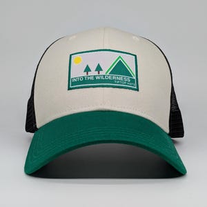 May include: A beige and green trucker hat with a black mesh back. The front panel has a rectangular patch with a mountain and tree design, the text "INTO THE WILDERNESS" and "TIPTOP HATS".