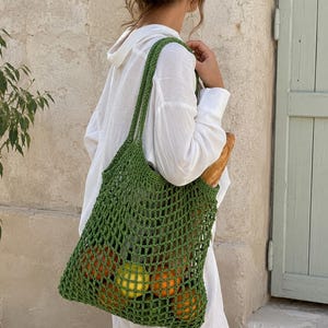 May include: A green crocheted net bag filled with oranges, lemons, and limes. The bag is carried over the shoulder. A loaf of bread is visible. The bag is made of a thick, open weave. The person is wearing a white shirt.