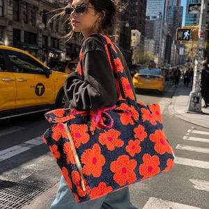 Può includere: Una grande borsa tote rettangolare realizzata in materiale testurizzato e soffice, caratterizzata da una base blu navy scuro con un motivo ripetuto di fiori arancioni brillanti con centri rosa vivaci. La borsa include due manici e una zip laterale.