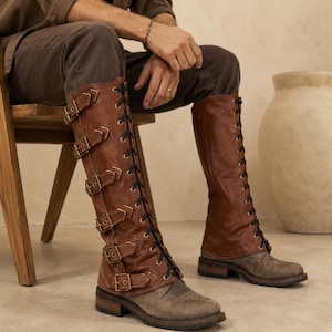 May include: Knee-high boots in a rich brown leather featuring a vertical row of five antique brass buckles on the outer side and black lace-up detailing running up the front. The boots have a low, stacked heel and a distressed grey toe cap.
