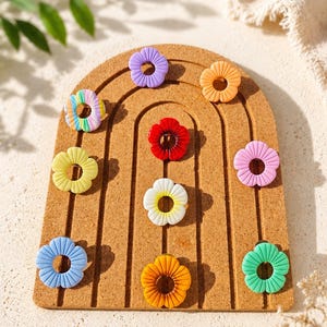 Peut inclure: Une collection de dix petits charms en forme de fleur circulaire dans des couleurs variées, notamment violet, orange, rouge, jaune, blanc, bleu, vert, rose et un motif rayé multicolore, disposés sur un panneau d'affichage en liège arqué.