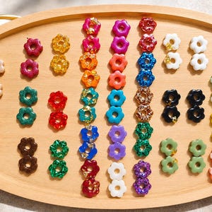 Peut inclure: Un plateau en bois présente un assortiment de perles en résine colorées en forme de fleurs. Les perles sont de différentes couleurs, dont le rose, l'orange, le bleu, le vert et le blanc, certaines contenant des paillettes dorées. Les perles sont disposées en rangées.