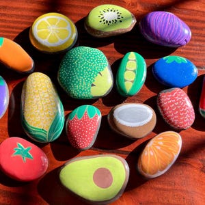 Può includere: Una collezione di sassi di fiume lisci dipinti a mano con vari frutti e verdure, tra cui un limone, kiwi, mais, fragola, pomodoro, avocado, arancia, melanzana, baccello di piselli e una fetta di anguria, disposti in un gruppo sparso.