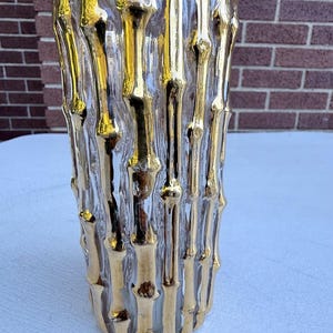 May include: A clear glass vase featuring a textured, raised gold-colored bamboo stalk design that wraps around the exterior. The cylindrical vessel has a smooth interior and a metallic finish on the vertical, segmented bamboo-like elements.