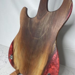 May include: A wooden electric guitar body with a dark brown finish and red resin accents. The guitar is displayed on a wooden stand, showcasing the back of the instrument. The body features a natural wood grain pattern.
