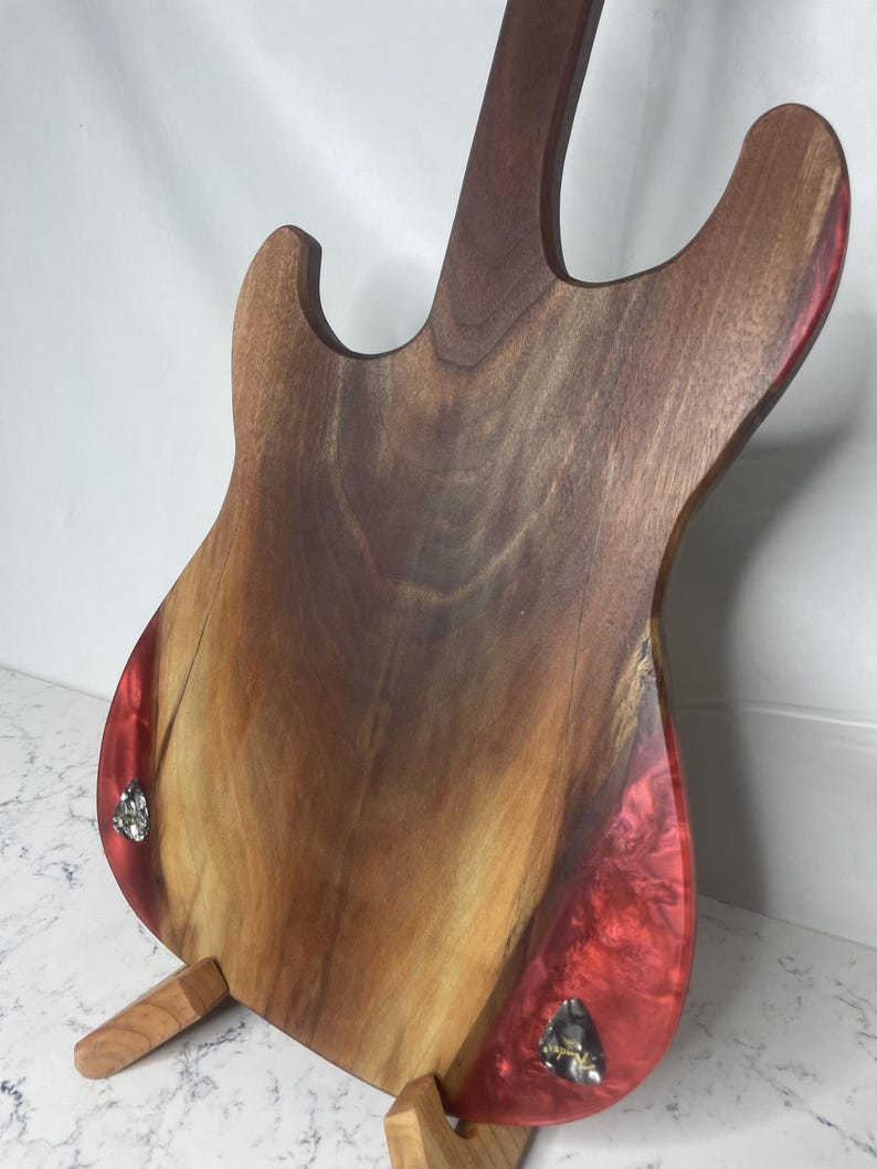 May include: A wooden electric guitar body with a dark brown finish and red resin accents. The guitar is displayed on a wooden stand, showcasing the back of the instrument. The body features a natural wood grain pattern.
