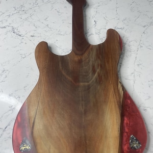 May include: A wooden guitar-shaped cutting board with a dark brown neck and body. The body has a reddish-brown hue with a red resin accent on the edges. Two guitar picks are embedded in the resin.